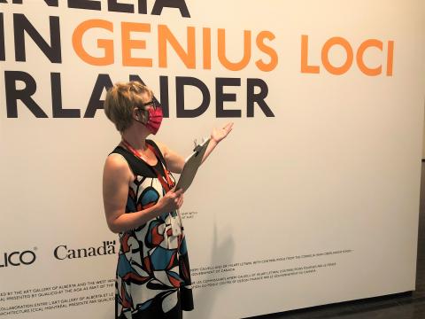 AGA Educator wearing a patterned tank dress with red mask standing in front of the title wall of Cornelia Oberlander: Genius Loci and is speaking about the late landscape architect. The exhibition is sponsored by Qualico and presented by the Poole Centre for Design.