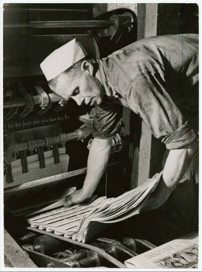 Black and white photo - Man picking up newspapers from printing machine